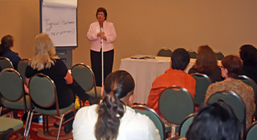 A woman stands in front of a room of people doing a presentation.