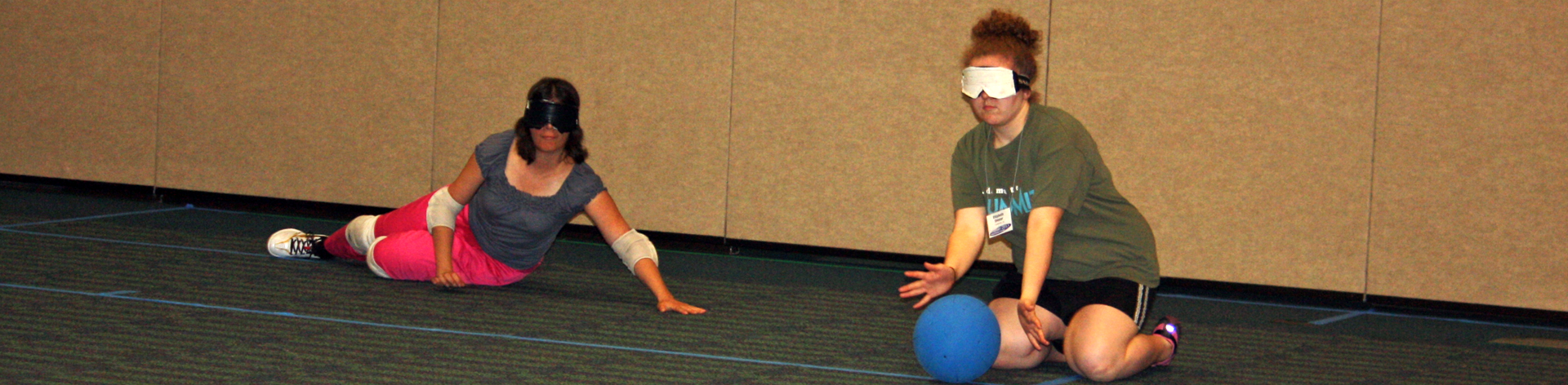 Two young members wearing sleepshades play goal ball. One reaches out to stop the ball from crossing into the goal.
